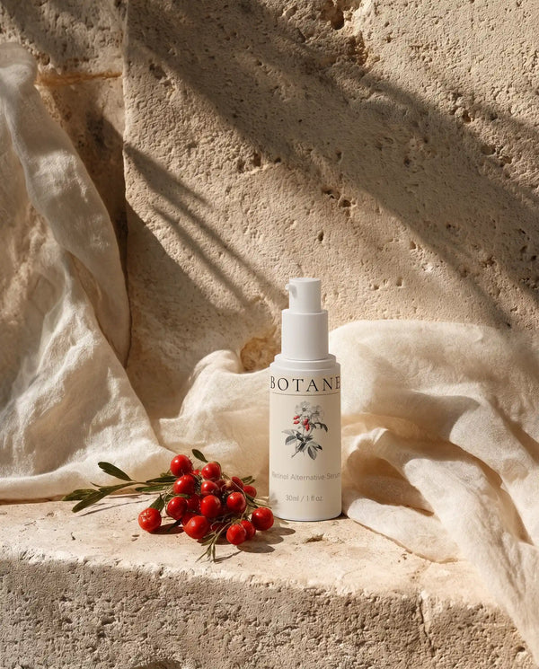 BOTANĒ Retinol Alternative Serum pump bottle on textured stone with red rosehips and draped linen in bright sunlight.
