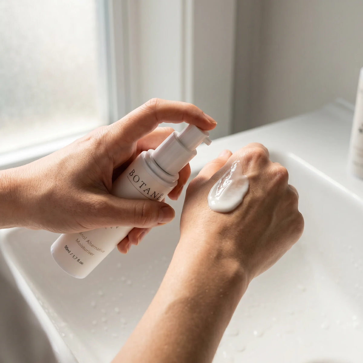 Hands dispensing BOTANĒ Retinol Alternative Moisturiser cream onto the back of a hand over a white bathroom sink.