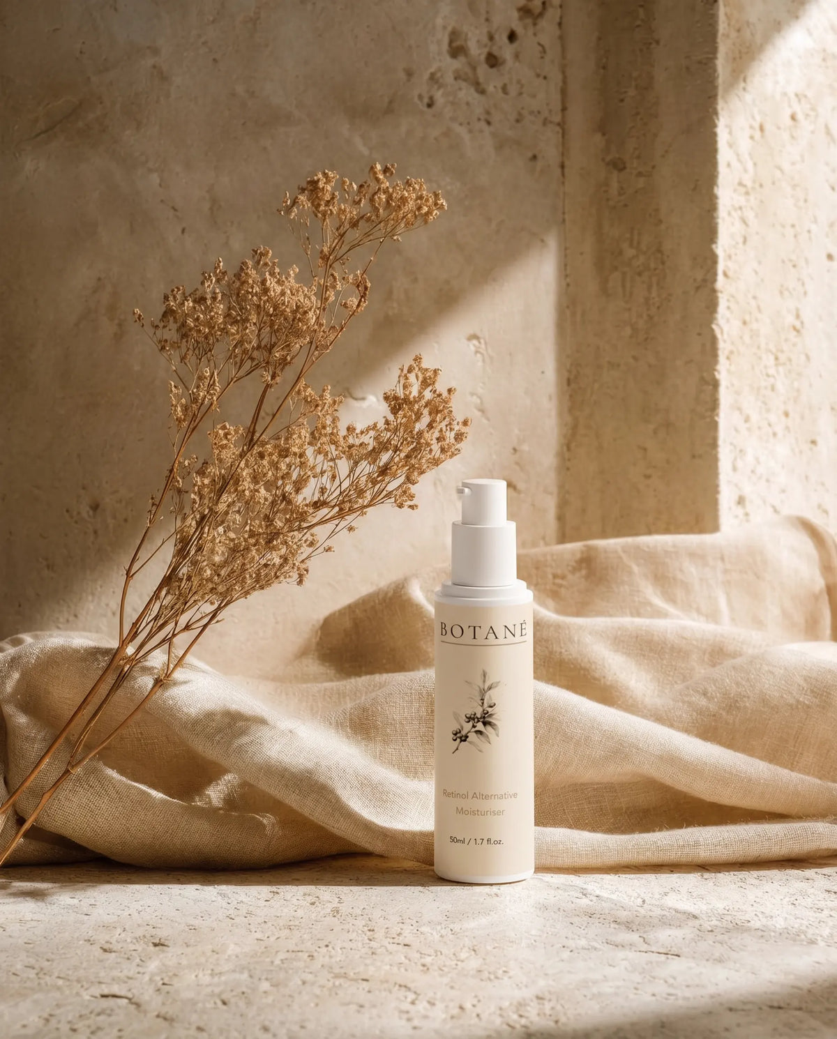 BOTANĒ Retinol Alternative Moisturiser pump bottle on textured stone with draped linen and dried flowers in warm sunlight.