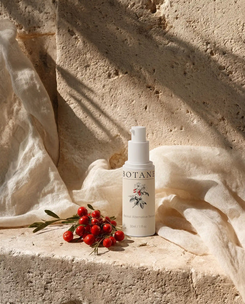 BOTANĒ Retinol Alternative Serum pump bottle on textured stone with red rosehips and draped linen in bright sunlight.